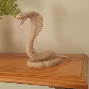 May include: A beige cobra statue with an open mouth, revealing fangs. The snake is posed in an upright position, resting on a textured base. The sculpture sits on a wooden shelf.