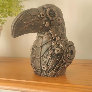 May include: A sculptural bust of a toucan bird rendered in a metallic dark grey finish. The surface features intricate mechanical details including visible rivets, circular gears, segmented plating, and coiled springs across the head and chest.