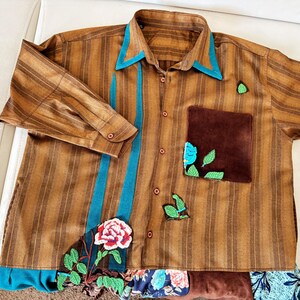 May include: A brown striped button-down shirt featuring a teal collar and vertical teal accents. The shirt includes a dark brown chest pocket, embroidered green leaf motifs, and a large embroidered rose at the bottom hem with floral fabric trim.