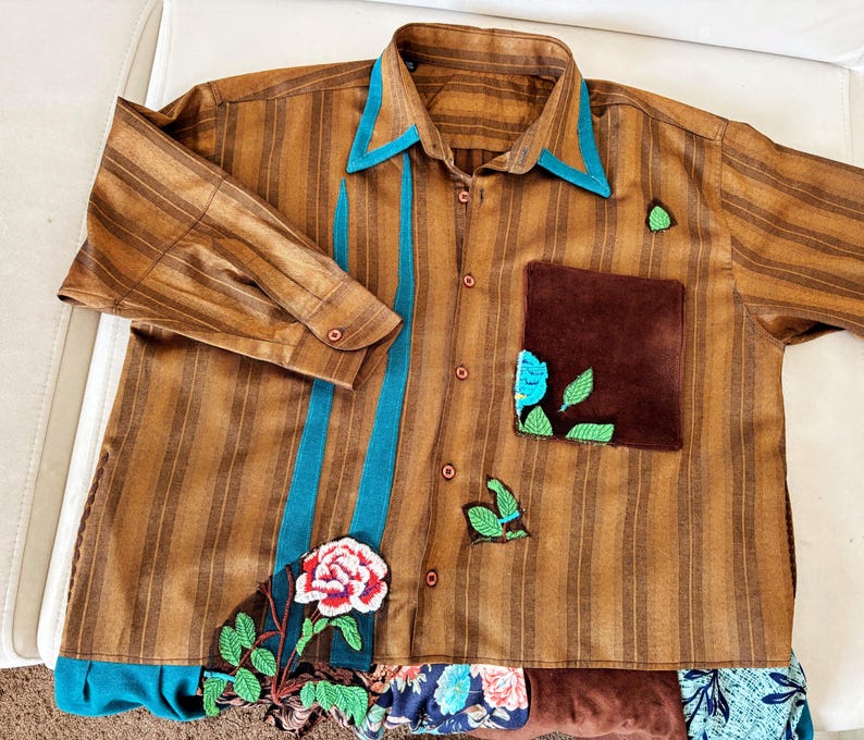 May include: A brown striped button-down shirt featuring a teal collar and vertical teal accents. The shirt includes a dark brown chest pocket, embroidered green leaf motifs, and a large embroidered rose at the bottom hem with floral fabric trim.