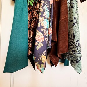 May include: Five hanging fabric swatches featuring a solid teal textile, a black and copper damask pattern, a navy blue floral print with roses, a solid brown suede-like material, and a light green fabric with a dark leaf and crosshatch pattern.