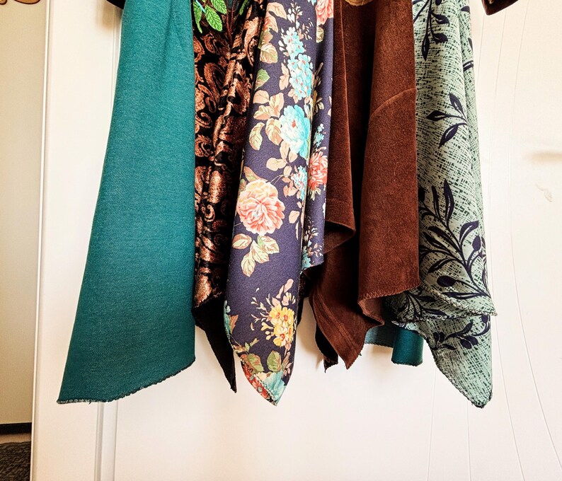 May include: Five hanging fabric swatches featuring a solid teal textile, a black and copper damask pattern, a navy blue floral print with roses, a solid brown suede-like material, and a light green fabric with a dark leaf and crosshatch pattern.
