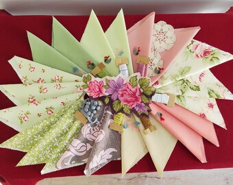 May include: A starburst arrangement of fabric triangles in various pastel colors and floral patterns. The center features a floral arrangement with spools of thread and decorative elements. The triangles are arranged on a red surface.