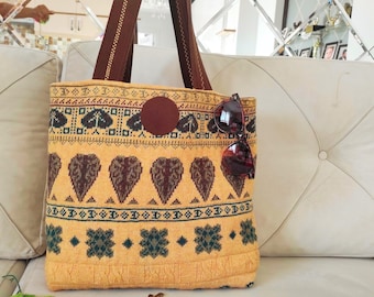 May include: A yellow tote bag featuring a brown leaf pattern, a circular brown patch, and dark brown straps. The bag is decorated with horizontal bands of geometric shapes and floral motifs in dark green and brown. A pair of sunglasses is attached.