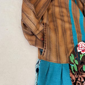 May include: A long-sleeved shirt featuring brown vertical stripes, a teal vertical panel, and a floral embroidery patch with a red and white rose and green leaves. The lower section includes a pleated teal fabric and a black patterned fabric panel.