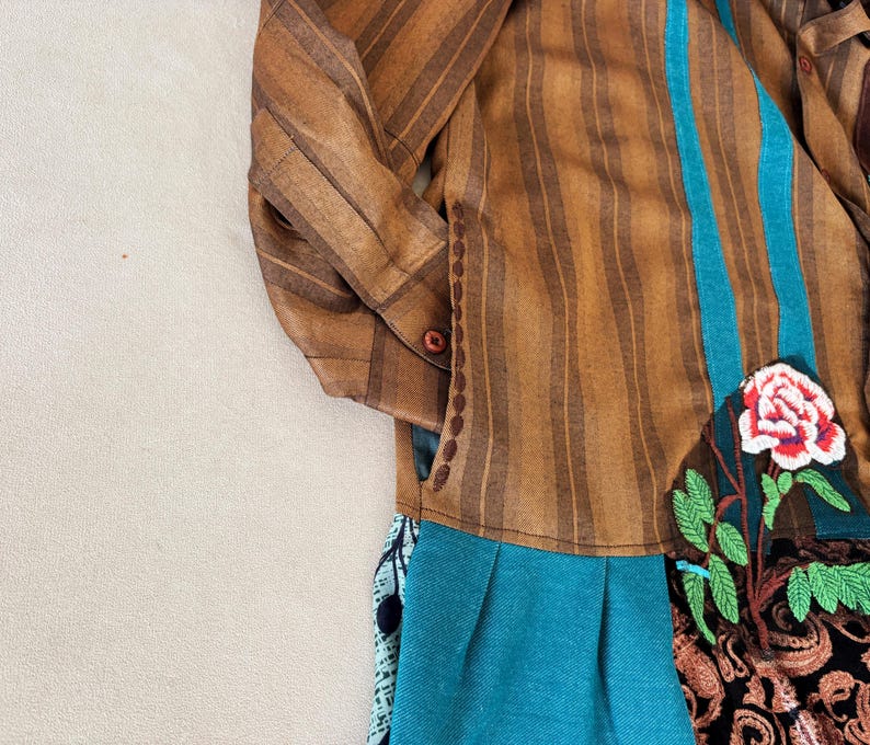 May include: A long-sleeved shirt featuring brown vertical stripes, a teal vertical panel, and a floral embroidery patch with a red and white rose and green leaves. The lower section includes a pleated teal fabric and a black patterned fabric panel.