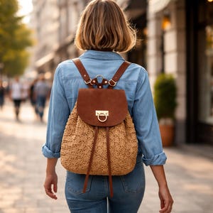 May include: A backpack featuring a textured, tan woven straw body and a smooth, dark brown leather flap. The flap is secured with a gold-toned metal ring clasp and features thin brown leather straps that hang down the front of the bag.