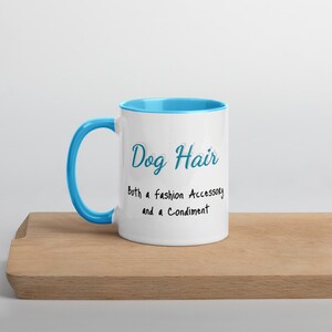 May include: White ceramic mug with a light blue rim and handle. The mug has black text that reads "Dog Hair both a fashion Accessory and a Condiment."