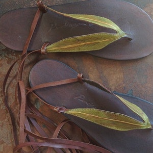 May include: Brown leather sandals with green leaf accents and long leather laces. The sandals have the brand name "Prada" written on the sole.