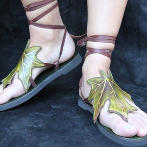 May include: A pair of brown leather sandals with green leaf accents. The sandals have laces that tie up the ankles.