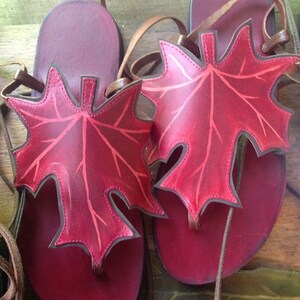 May include: A pair of red leather sandals with maple leaf cutouts. The sandals have brown leather straps and are tied at the ankle.