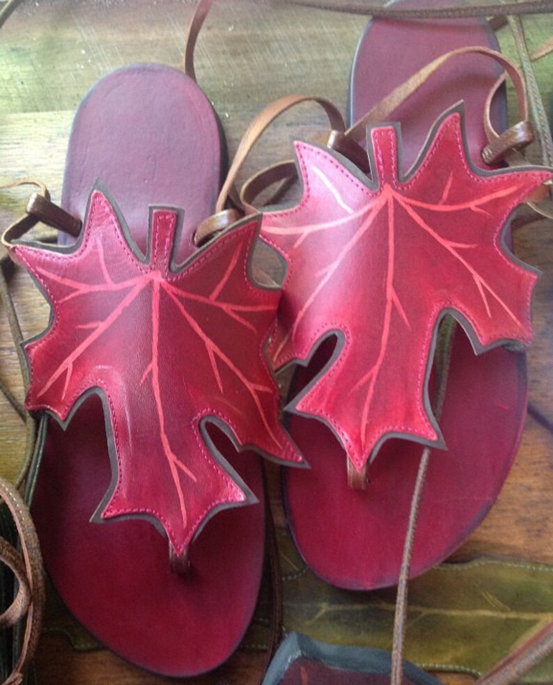 May include: A pair of red leather sandals with maple leaf cutouts. The sandals have brown leather straps and are tied at the ankle.