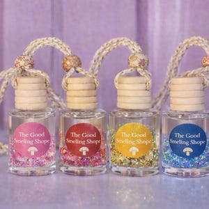 May include: Four small, clear glass bottles with wooden lids and decorative cords. Each bottle has a colorful label with the text "The Good Smelling Shope" and a mushroom graphic. The bottles are filled with glitter.