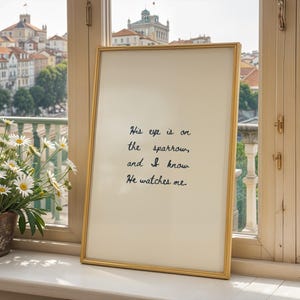 Puede incluir: Un marco dorado que contiene una impresión con un fondo crema y texto manuscrito azul oscuro: "His eye is on the sparrow, and I know He watches me". El marco está apoyado cerca de una ventana.