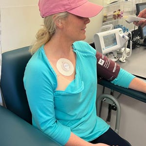 May include: A woman wearing a light blue long-sleeved shirt and a pink baseball cap. A medical device is attached to her chest. A blood pressure cuff is on her arm. She is seated in a blue chair.