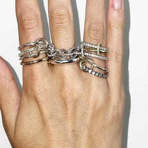 May include: A hand displaying a collection of rings in various styles. The rings include gold and silver bands, some adorned with small, sparkling stones. The designs range from simple bands to more intricate, knot-like structures, showcasing a mix of textures and finishes.