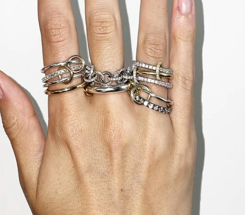 K&ouml;nnte beinhalten: Eine Hand, die eine Sammlung von Ringen in verschiedenen Stilen zeigt. Die Ringe umfassen goldene und silberne B&auml;nder, einige mit kleinen, funkelnden Steinen verziert. Die Designs reichen von einfachen B&auml;ndern bis hin zu komplizierteren, knotenartigen Strukturen, die eine Mischung aus Texturen und Oberfl&auml;chen zeigen.