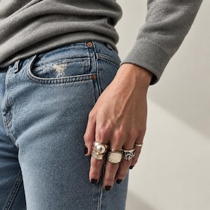 May include: A collection of four silver-toned rings worn on the fingers. One ring features a round, polished metallic center, another has a smooth, square-shaped face, and two others display intricate, textured, or twisted metal band designs.