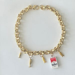 Könnte beinhalten: Eine goldfarbene Gliederkette mit strukturierter Oberfläche und Karabinerverschluss. Die Kette ist mit vier kleinen, goldfarbenen Fisch-Anhängern und einem rechteckigen Miniatur-Anhänger verziert, der eine Packung Marlboro Medium 100s zeigt.