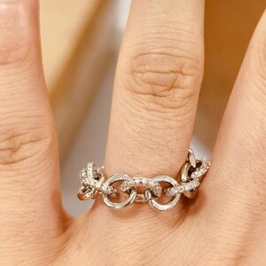 K&ouml;nnte beinhalten: Ein silberner Ring mit einem Kettenglied-Design, besetzt mit kleinen, funkelnden Steinen. Der Ring wird an einem Finger getragen und zeigt seine komplizierten Details und die reflektierende Oberfl&auml;che. Das Schmuckst&uuml;ck ist ein Modeaccessoire.