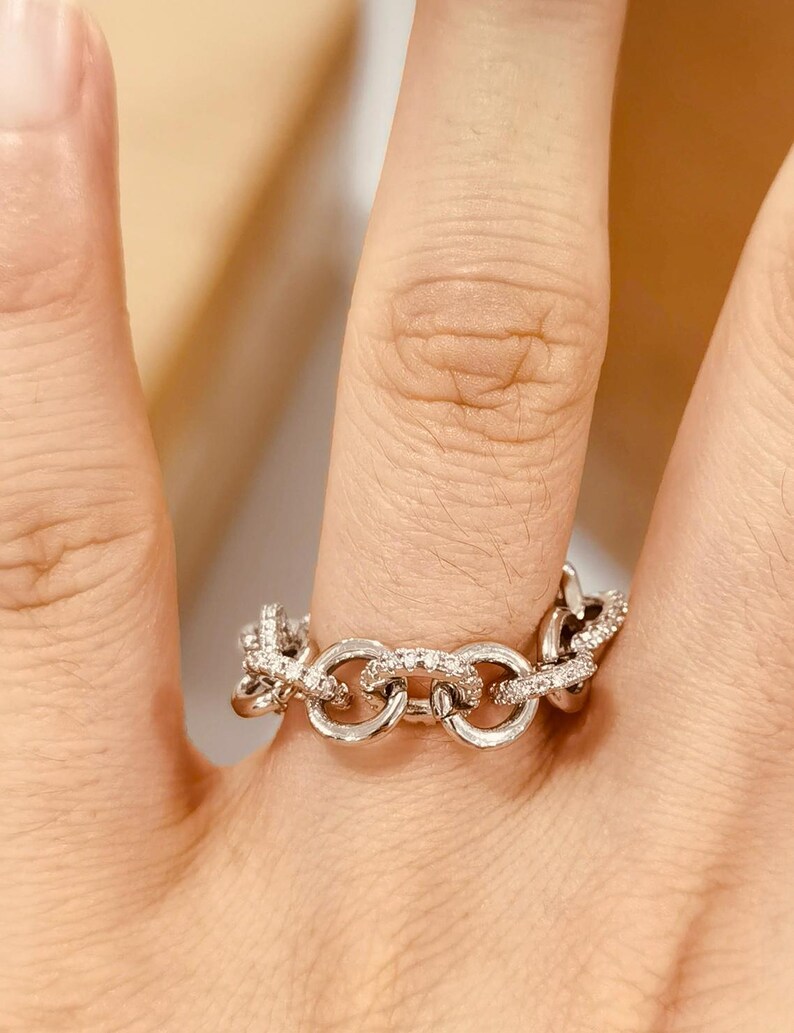 K&ouml;nnte beinhalten: Ein silberner Ring mit einem Kettenglied-Design, besetzt mit kleinen, funkelnden Steinen. Der Ring wird an einem Finger getragen und zeigt seine komplizierten Details und die reflektierende Oberfl&auml;che. Das Schmuckst&uuml;ck ist ein Modeaccessoire.