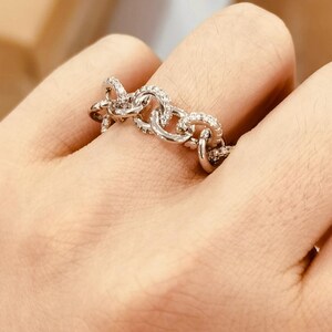 K&ouml;nnte beinhalten: Ein silberfarbener Ring mit einem Kettenglied-Design. Der Ring ist mit kleinen, klaren Edelsteinen besetzt. Der Ring wird an einem Finger getragen.