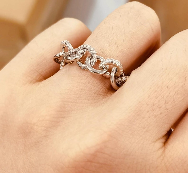 K&ouml;nnte beinhalten: Ein silberfarbener Ring mit einem Kettenglied-Design. Der Ring ist mit kleinen, klaren Edelsteinen besetzt. Der Ring wird an einem Finger getragen.
