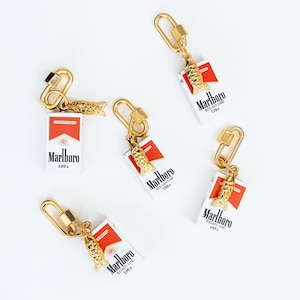 Peut inclure: Cinq porte-clés composés d'une réplique miniature de paquet de cigarettes Marlboro blanc et rouge, attachée à un fermoir mousqueton doré et à une petite breloque poisson dorée texturée. Chaque paquet affiche le logo Marlboro et du texte numérique.