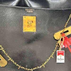 Könnte beinhalten: Schwarze Lederhandtasche mit goldfarbener Hardware, einschließlich Verschluss und Kette. Ein kleiner Marlboro-Zigarettenpackungs-Anhänger hängt an der Kette. Die Tasche trägt den Schriftzug "HERMES PARIS".