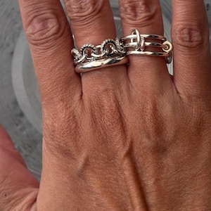 K&ouml;nnte beinhalten: Nahaufnahme von zwei silbernen Ringen an einem Finger. Ein Ring hat ein Kettenmuster mit kleinen, klaren Steinen. Der andere Ring hat ein einzigartiges, geometrisches Design mit mehreren B&auml;ndern und einem kreisf&ouml;rmigen Element. Die Ringe sind gl&auml;nzend und reflektieren das Licht.