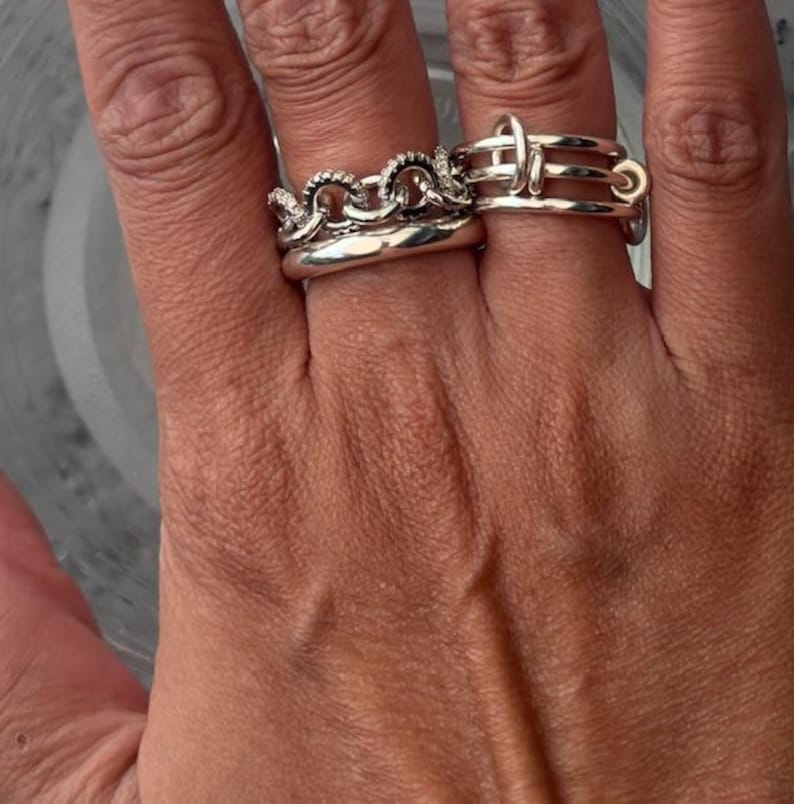 K&ouml;nnte beinhalten: Nahaufnahme von zwei silbernen Ringen an einem Finger. Ein Ring hat ein Kettenmuster mit kleinen, klaren Steinen. Der andere Ring hat ein einzigartiges, geometrisches Design mit mehreren B&auml;ndern und einem kreisf&ouml;rmigen Element. Die Ringe sind gl&auml;nzend und reflektieren das Licht.