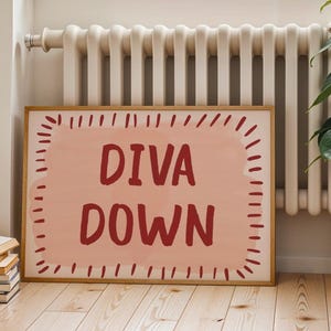 May include: A framed art print with the words "DIVA DOWN" in a bold, red font. The text is set against a light pink background with a dotted border, encased in a wooden frame. The artwork is leaning against a radiator.