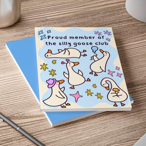 May include: A notebook with a light blue cover and a cream-coloured cover featuring cartoon geese and the text "Proud member of the silly goose club". The geese are in various poses with stars and other decorative elements.