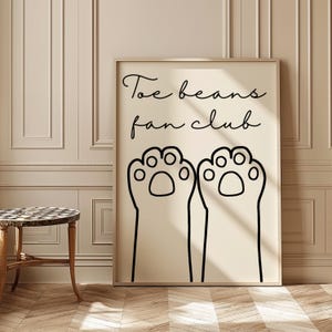 Peut inclure: Une affiche beige avec du texte noir et des illustrations. Le texte dit "Toe beans fan club". Deux empreintes de pattes noires sont sous le texte.