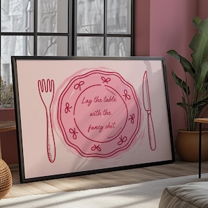 May include: Framed art print featuring a pink plate, fork, and knife illustration. The plate has the text "Lay the table with the fancy shit" in a handwritten style. The artwork is framed in a black frame.