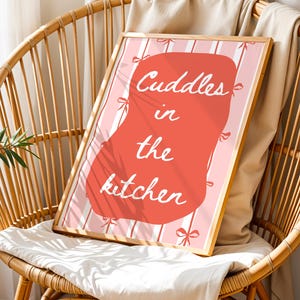 Op de afbeelding: Een ingelijste kunstdruk met de tekst "Cuddles in the kitchen" in witte letters op een koraalkleurige vorm, tegen een roze en witte gestreepte achtergrond. De print wordt gepresenteerd op een houten stoel met een wit kussen.