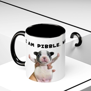 May include: White ceramic mug with a black interior and handle. The mug features the text "I AM PIBBLE. WA" and a graphic of a light-colored puppy with its arms outstretched. The puppy has a black and grey head.