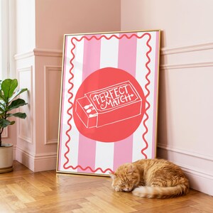 May include: A rectangular wall art print in a gold frame featuring a pink and white vertical striped background with a wavy red border. Centered is a large red circle containing a white line drawing of a matchbox with the text PERFECT MATCH.