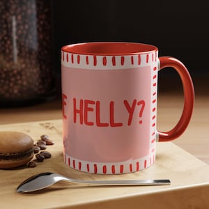 May include: A red-handled coffee mug with the text "ARE HELLY?" in red on a pink background. The mug has a red interior and is decorated with a white and red dotted border. A spoon and a macaron are on a wooden surface.