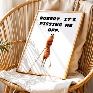 Puede incluir: Una impresión artística enmarcada con una ilustración de dibujos animados de una figura naranja con piernas largas. La impresión muestra el texto "ROBERT. IT'S PISSING ME OFF." en negro. La obra de arte está enmarcada en un marco de madera y descansa sobre una silla de mimbre.