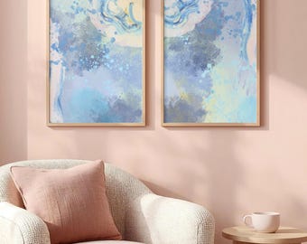 May include: Two rectangular wall art prints in light wooden frames featuring abstract watercolor designs in shades of blue, pale yellow, and white with paint splatters, swirling brushstrokes, and textured gradients on a light background.
