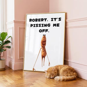 May include: A framed wall art print featuring a white background with the black text "ROBERT. IT'S PISSING ME OFF." above a stylised, tan, elongated creature with thin limbs and a small head, presented in a thin gold-coloured metal frame.
