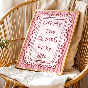 Könnte beinhalten: Ein gerahmter Druck mit dem Text "Off My Tits On M&S Picky Bits". Der Druck hat einen rosa-weiß gestreiften Hintergrund und einen dekorativen Rand. Der Rahmen ist helles Holz, und der Druck wird auf einem Korbstuhl präsentiert.