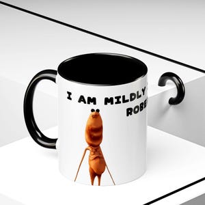 May include: White ceramic mug with a black interior and handle. The mug features a cartoon character and the text "I AM MILDLY ROBE". The character is a brown, humanoid figure with thin limbs and small eyes.