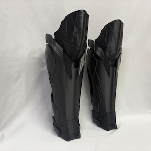 Pode incluir: Um par de caneleiras preto mate com um design estruturado de várias camadas, painéis angulares e cristas elevadas. Estas peças de proteção para as pernas têm uma silhueta alta e cónica com um acabamento liso e detalhes geométricos.