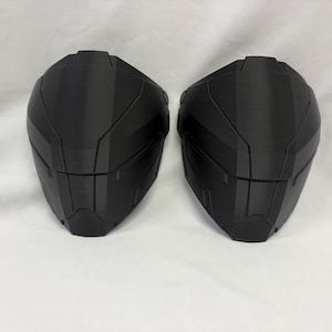 Könnte beinhalten: Zwei schwarze, kantige, futuristisch aussehende Helmhälften. Die Helme haben ein segmentiertes Design mit mehreren Ebenen und Rippen, was auf eine High-Tech- oder Science-Fiction-Ästhetik hindeutet. Sie sind wahrscheinlich 3D-gedruckt.