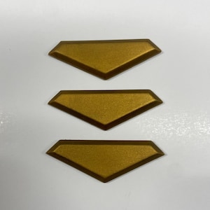 May include: Three identical gold-colored, trapezoidal-shaped decorative pieces arranged in a vertical column. Each piece features a flat, textured surface with beveled edges that taper to a point at the bottom, creating a distinct geometric shape.