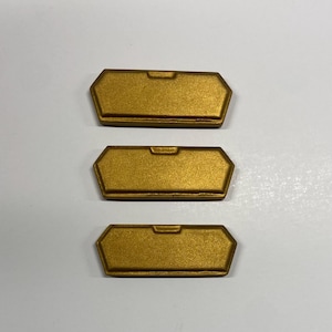 Peut inclure: Trois plaques métalliques rectangulaires identiques de couleur dorée avec des bords biseautés et une petite encoche centrée sur le bord supérieur. Chaque plaque présente une finition mate lisse et une forme hexagonale allongée uniforme.