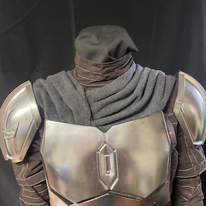 Può includere: Un'armatura pettorale e spallacci color argento con un cappuccio e una copertura per il collo grigio scuro. Il costume ha un design a strati con cuciture visibili e una copertura per la testa nera. L'armatura ha una lucentezza metallica.