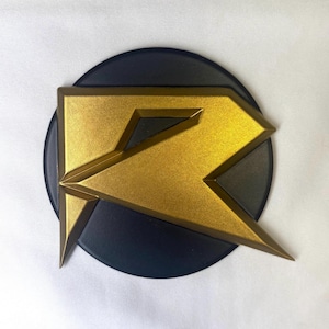 Könnte beinhalten: Ein kreisförmiges schwarzes Emblem mit einem stilisierten goldenen Buchstaben R mit scharfen, kantigen Linien. Das goldene Element hat eine texturierte, metallische Oberfläche, während die runde Basis mattschwarz ist.